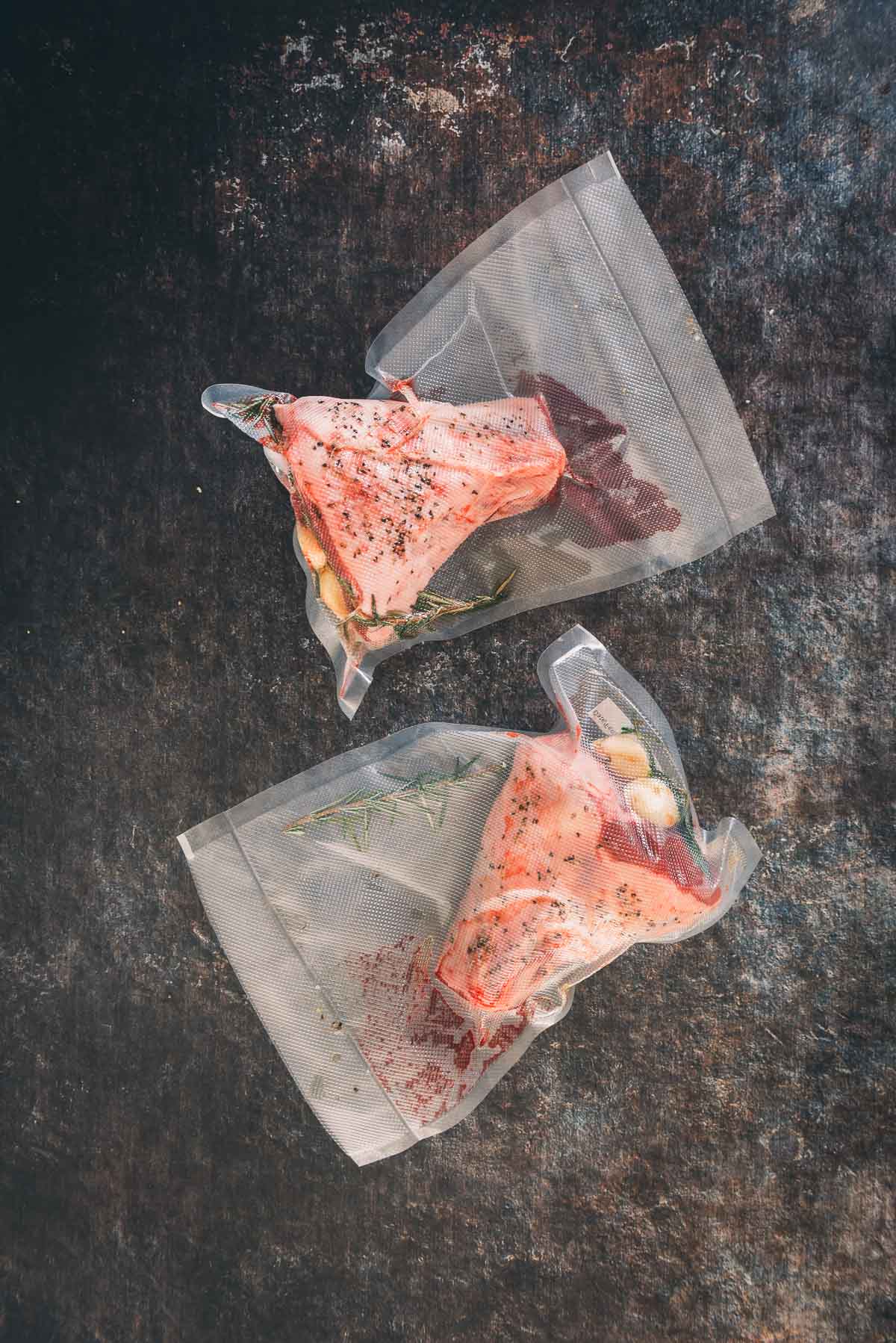 Two lamb shanks in vacuum sealed bags on a dark background.
