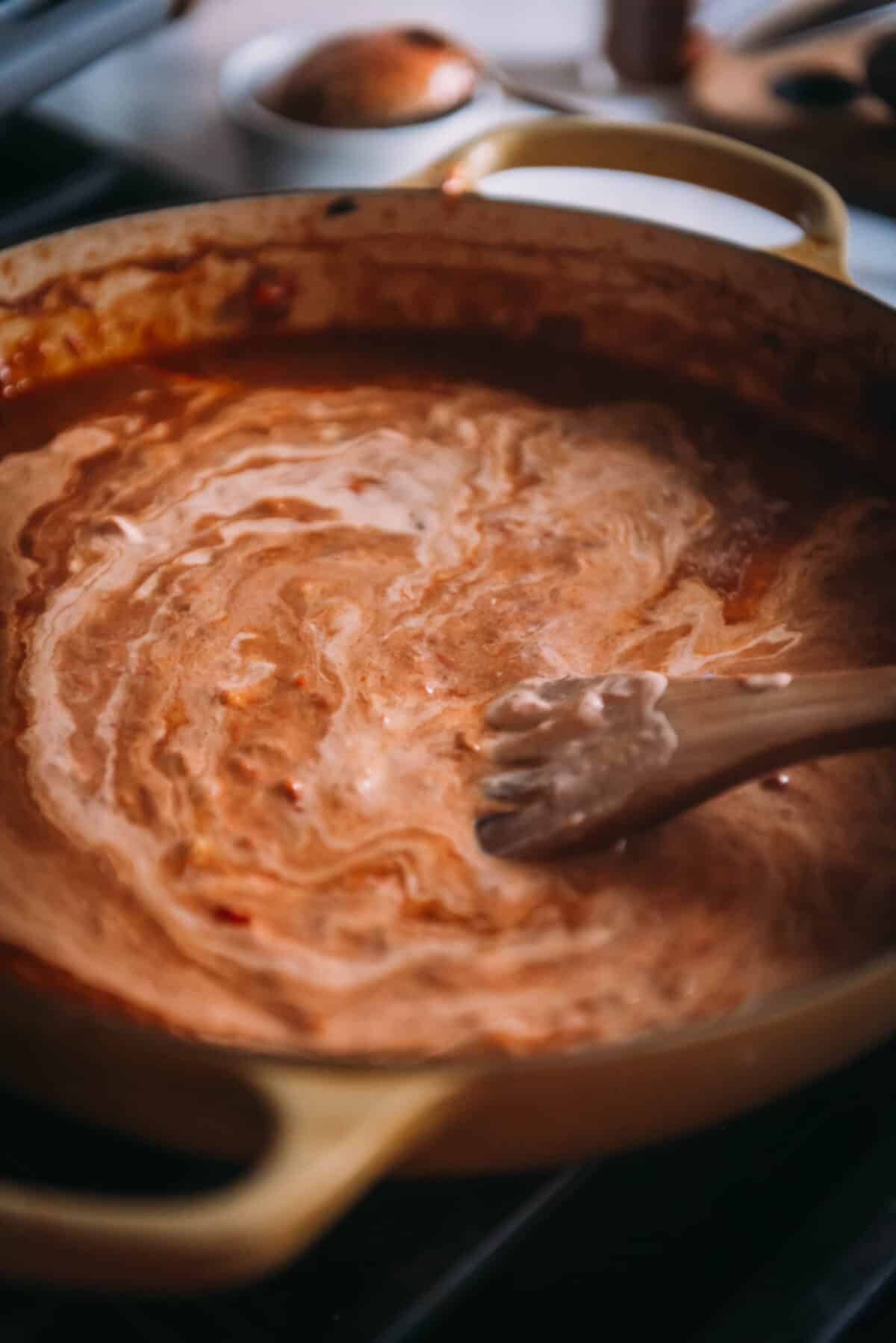 A pot of sauce with swirls of sour cream on the stove with a wooden spoon.