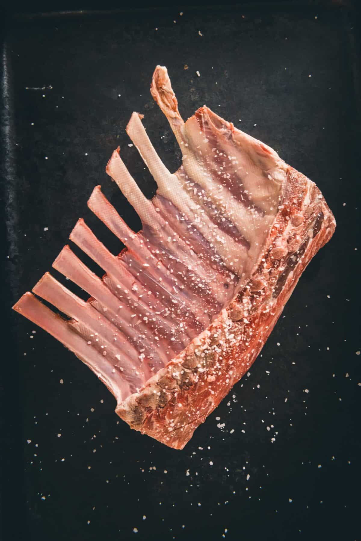 A salted lamb rack on a black tray.