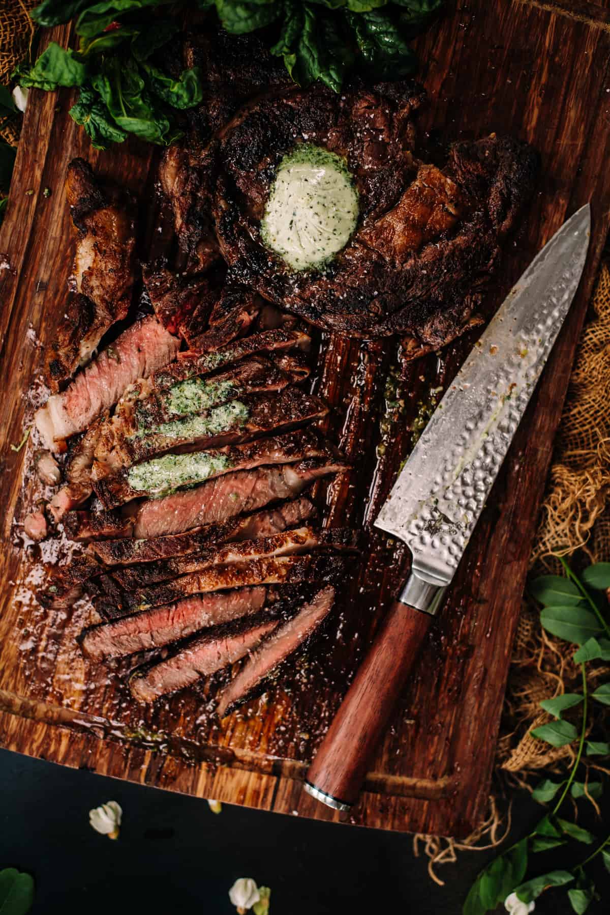 A steak on a cutting board with herbs and a knife.