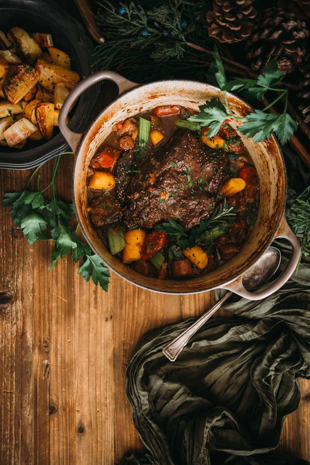 An overhead shot of a Dutch oven with a pot roast and vegetables on a wooden table.