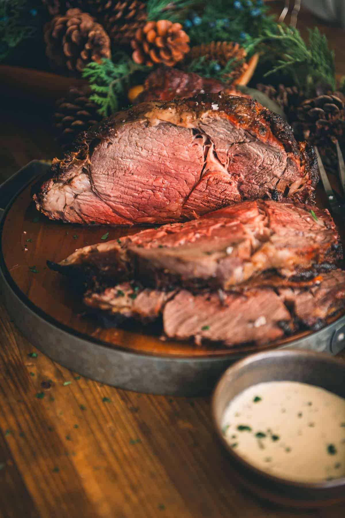 A standing rib roast with sauce and pine cones on a wooden table.