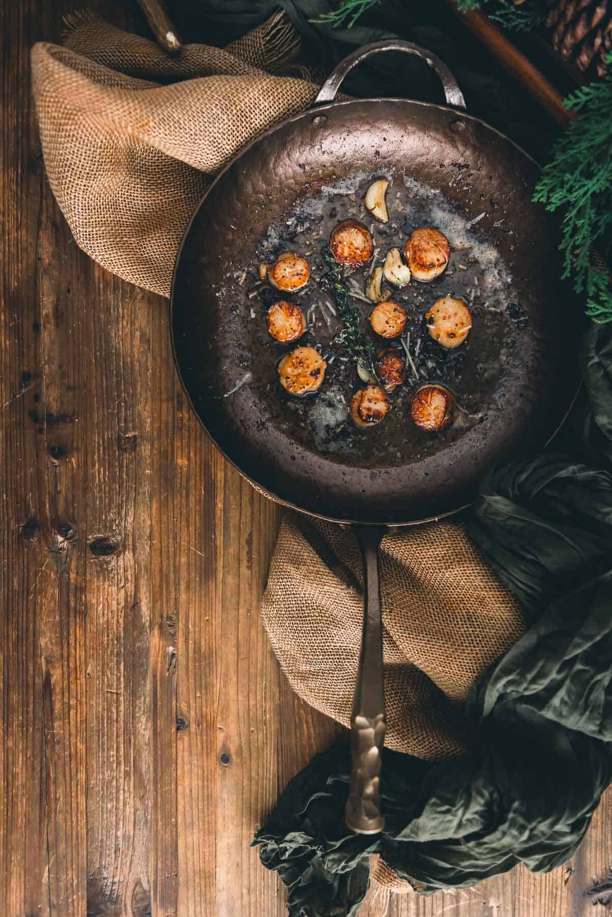A frying pan with scallops and pine cones on a wooden table.