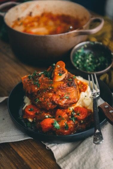 Braised pork shanks, with the bone sticking out on a platter with carrots and potatoes garnished with parsley on a table.