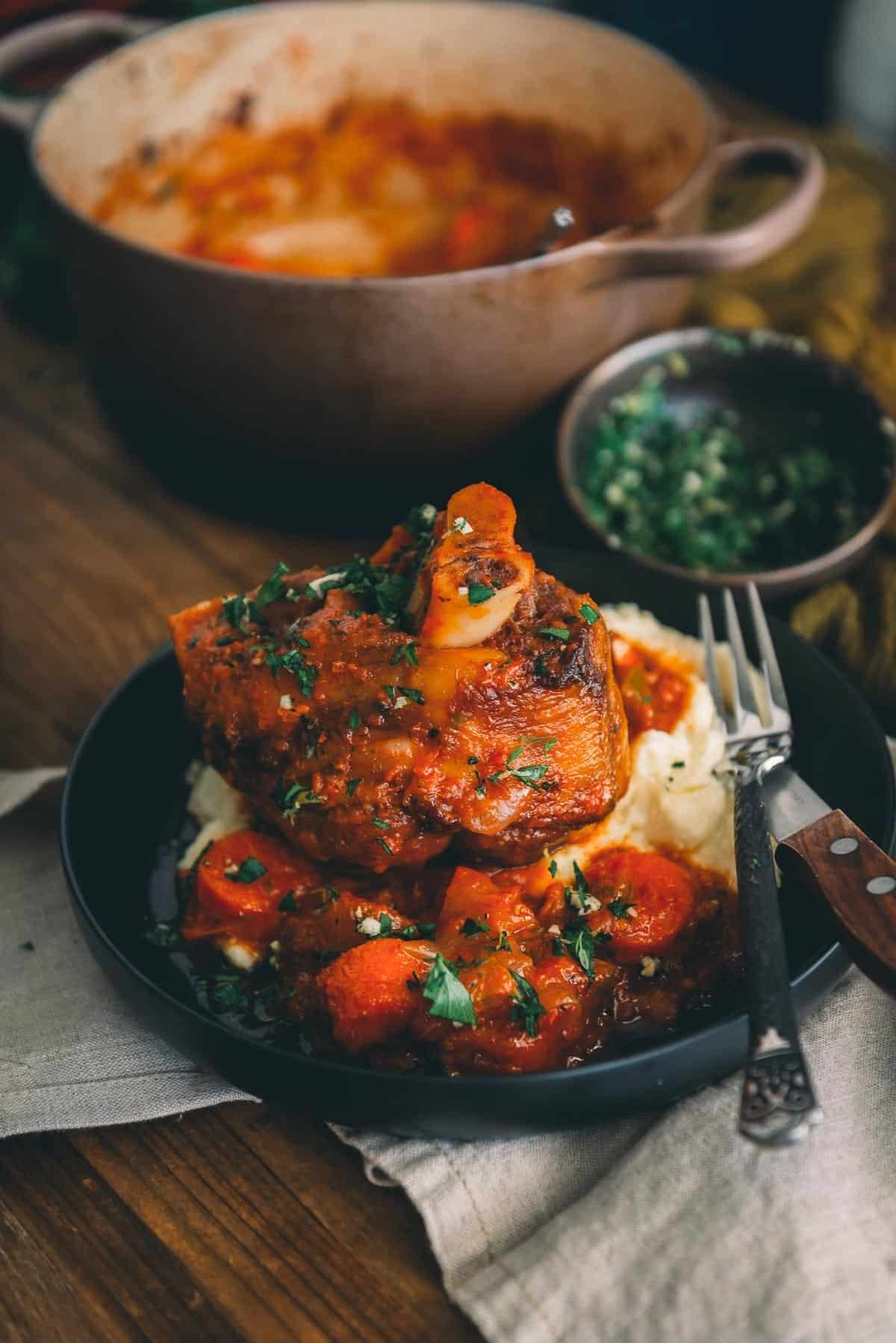 Braised pork shanks, with the bone sticking out on a platter with carrots and potatoes garnished with parsley on a table.