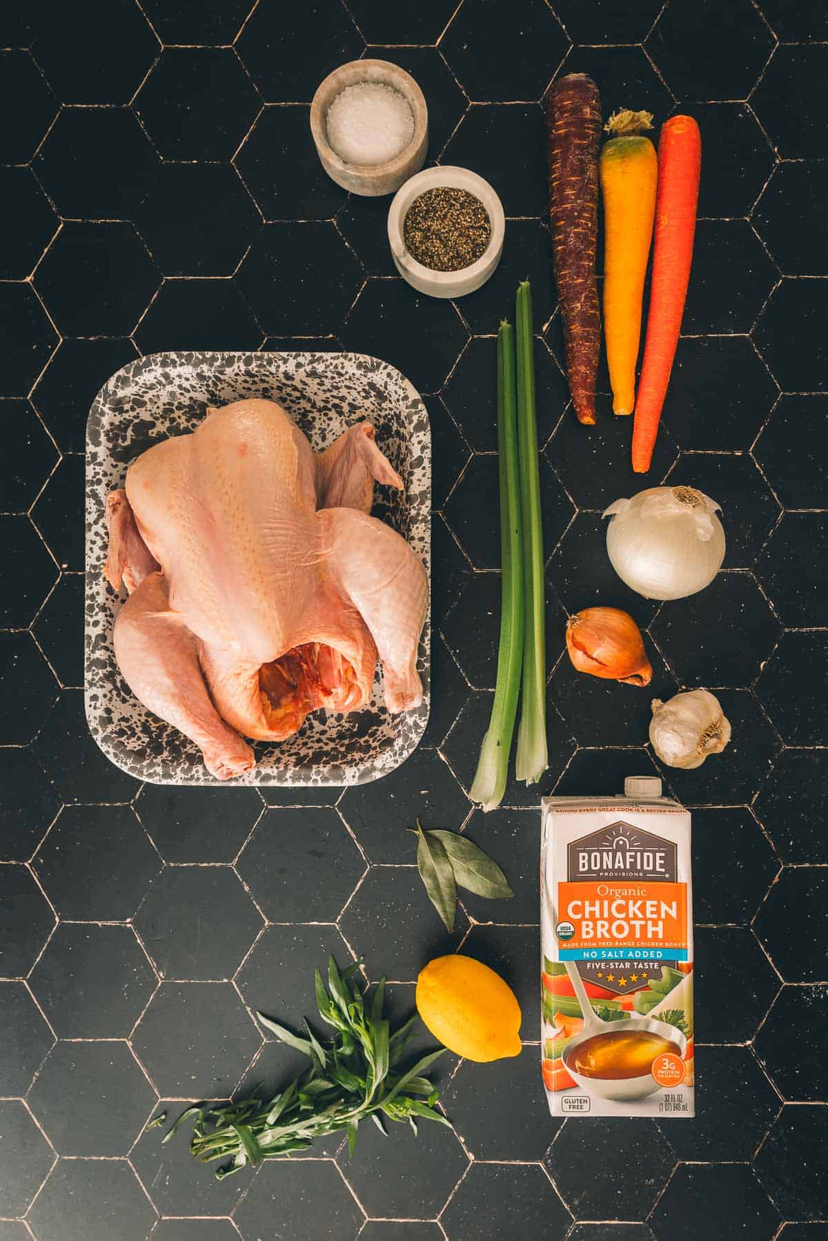 A whole chicken, carrots, celery and other ingredients on a black countertop.