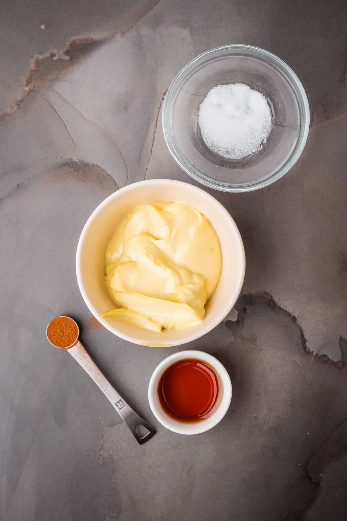 A bowl of homemade mayo with other ingredients and a spoon on a gray surface.