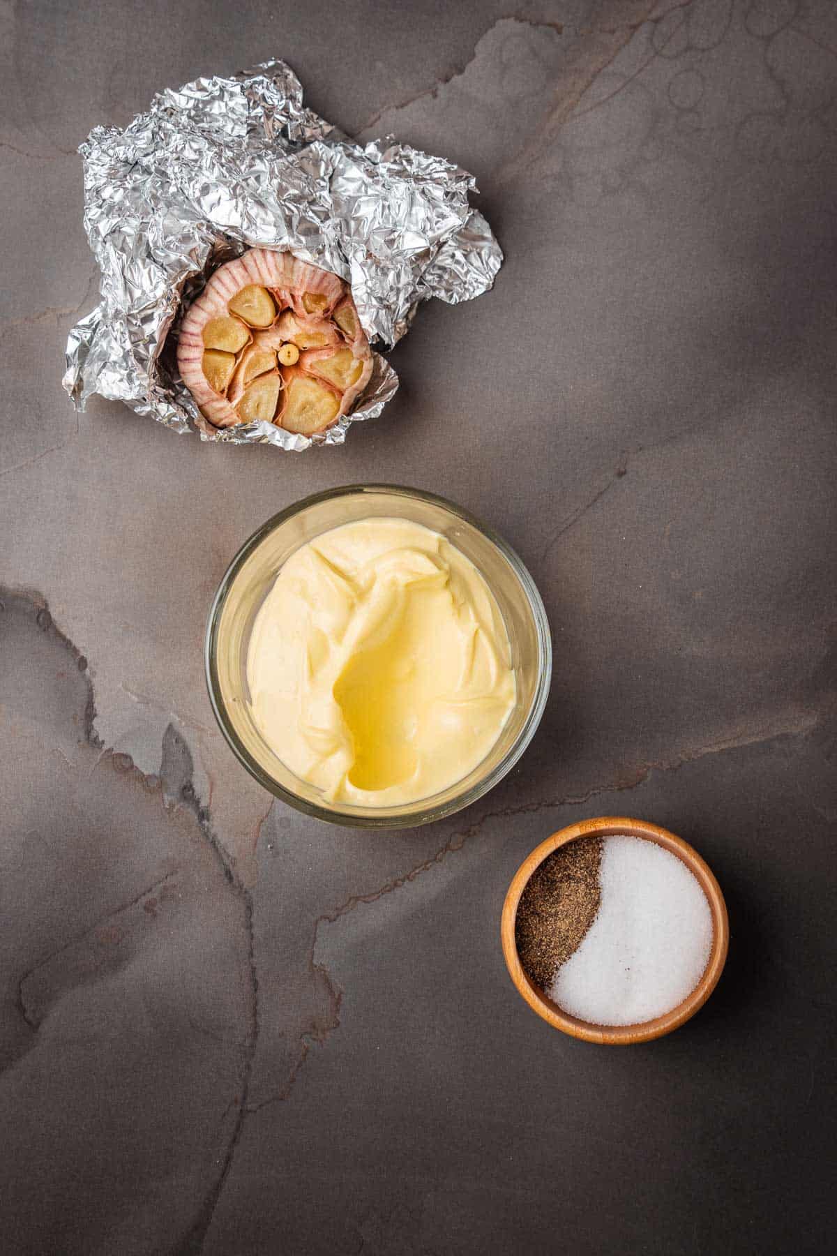 A bowl of aioli, roasted garlic, and a bowl of salt and pepper on a gray background.