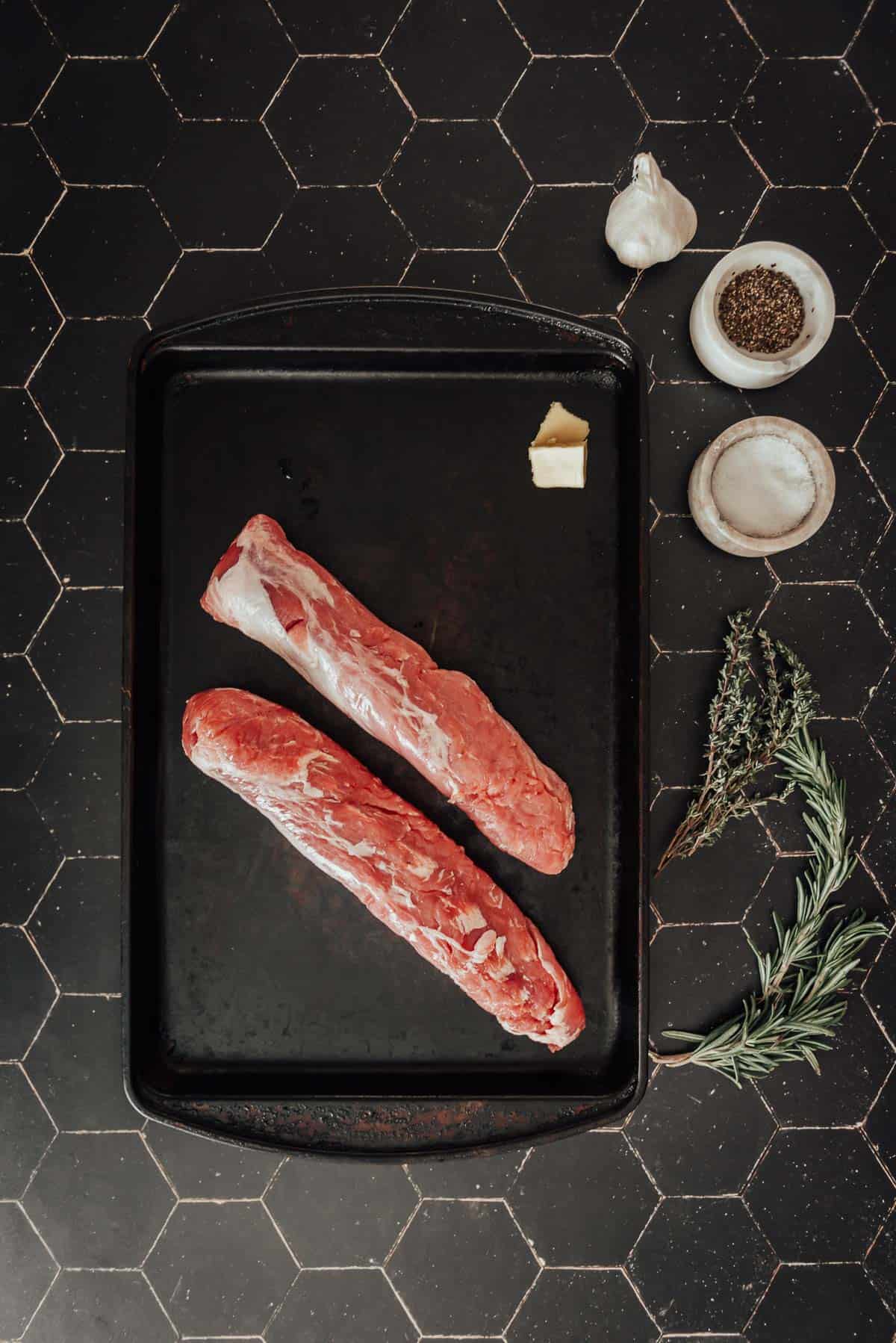 Two pork tenderloins on a tray with herbs and spices.