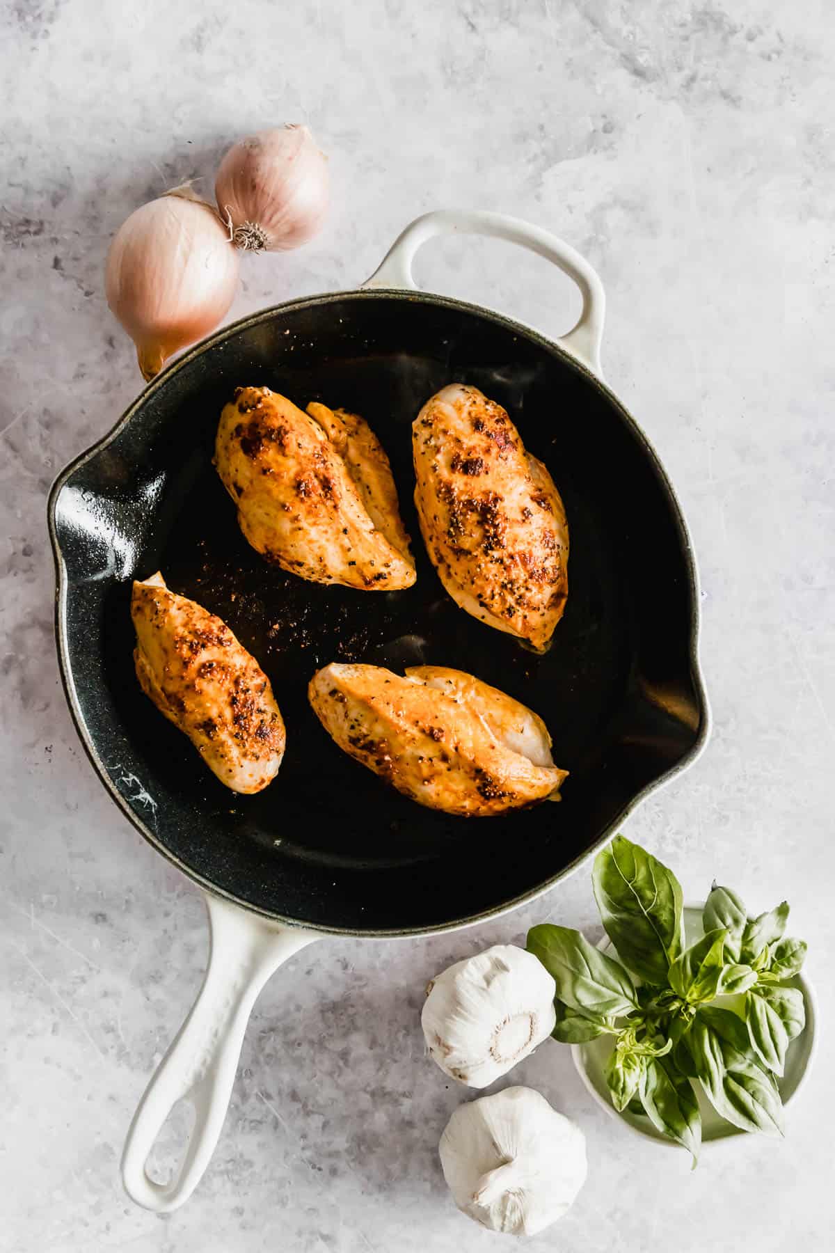 Four seasoned chicken breasts in a cast iron skillet, surrounded by garlic, basil, and onions on a marble surface.