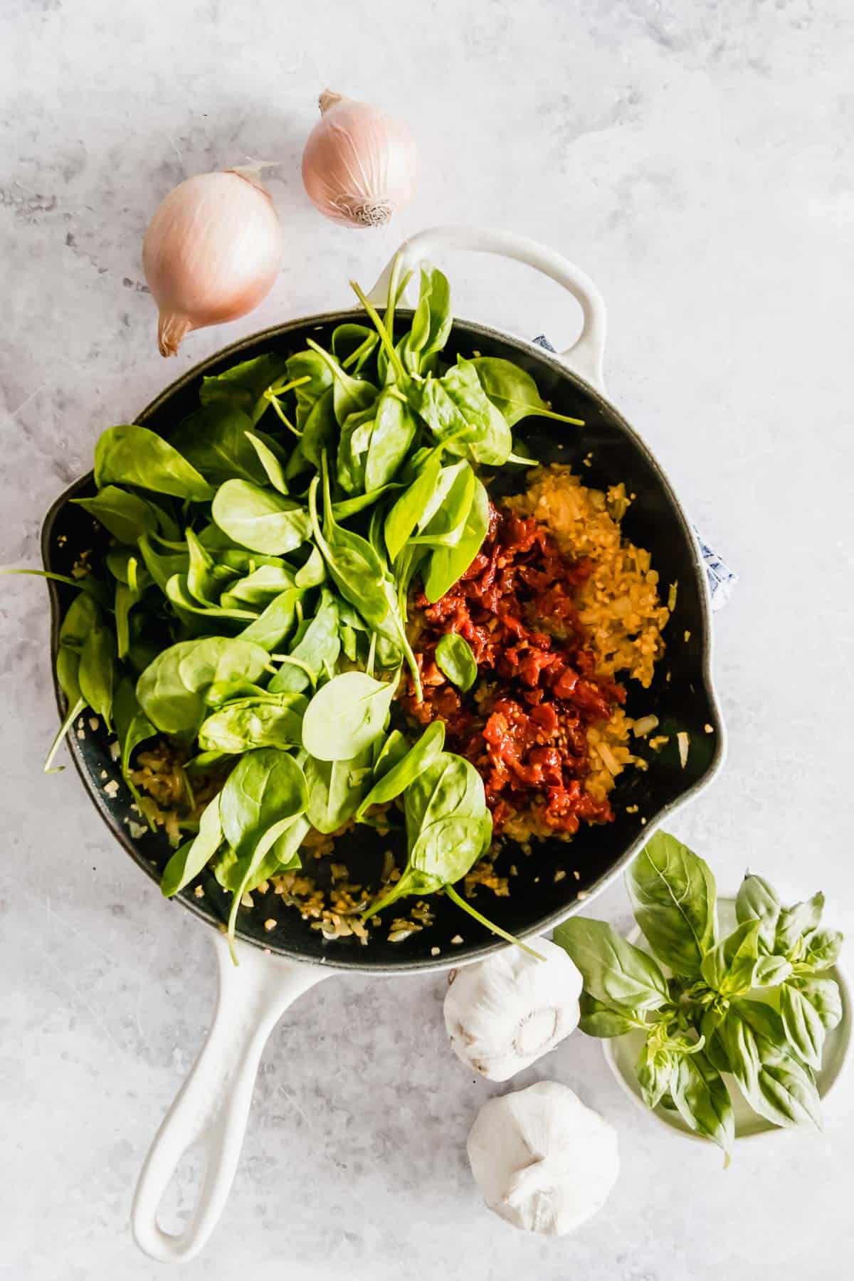 A skillet filled with rice, sun-dried tomatoes, and fresh spinach, surrounded by garlic and onions on a marble countertop.