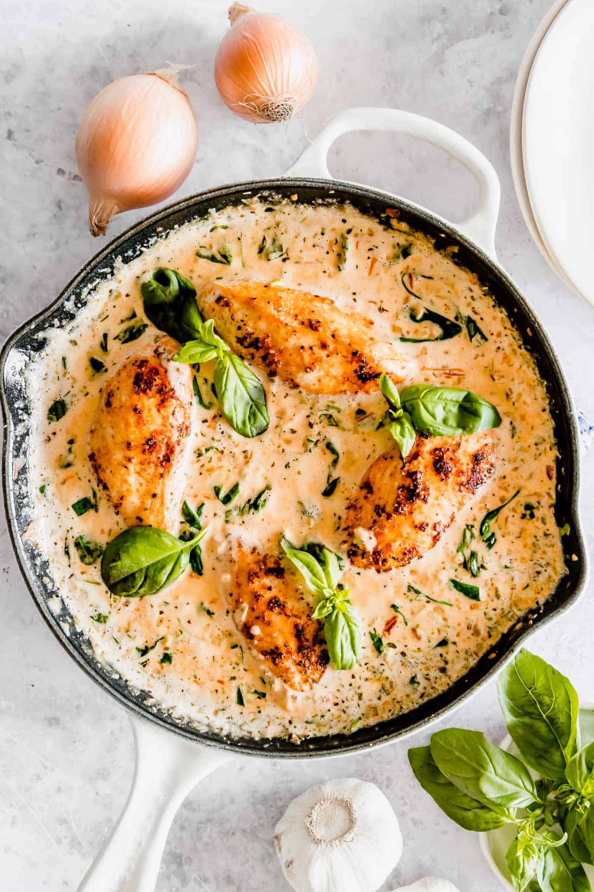 Four pieces of chicken in a creamy sauce in a skillet, garnished with basil, surrounded by onions and garlic on a marble surface.