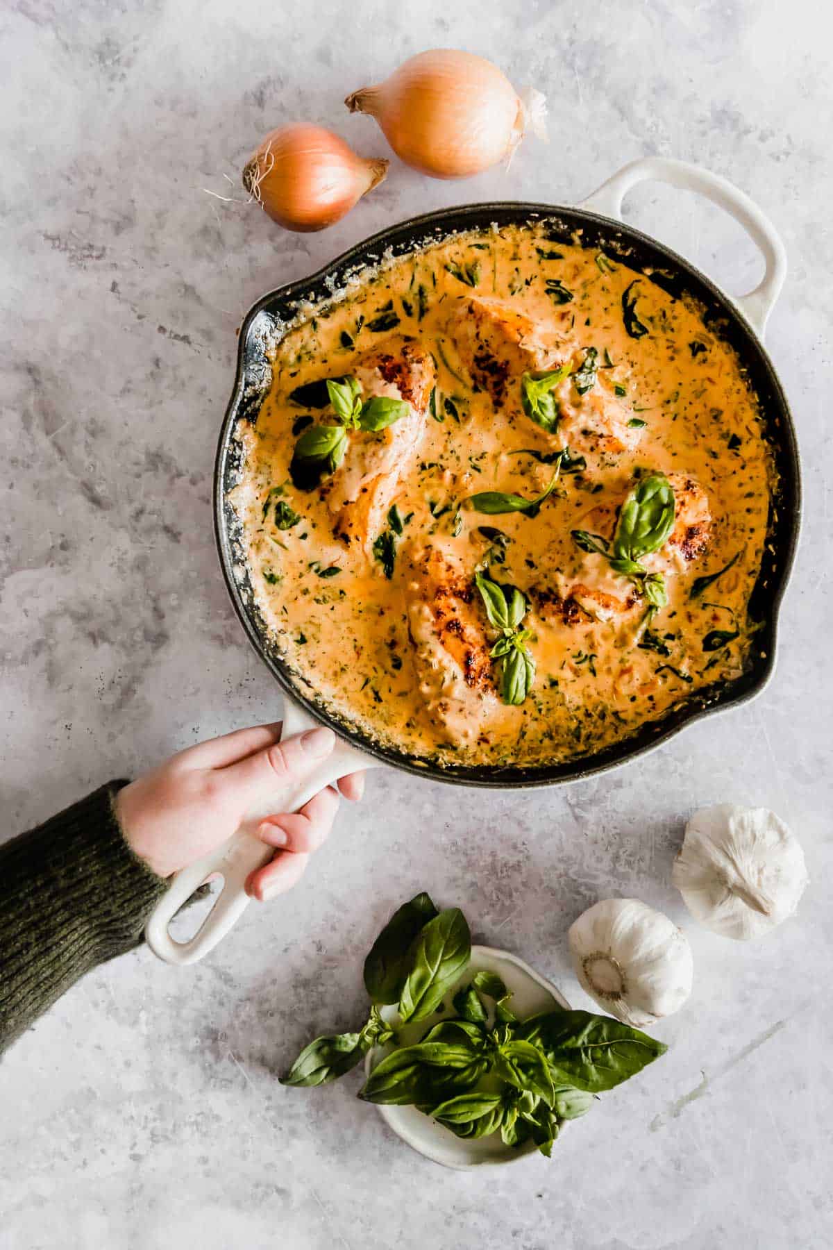 A person's hand holding a skillet containing creamy tuscan chicken with fresh basil on top, surrounded by raw garlic and onions on a marble surface.