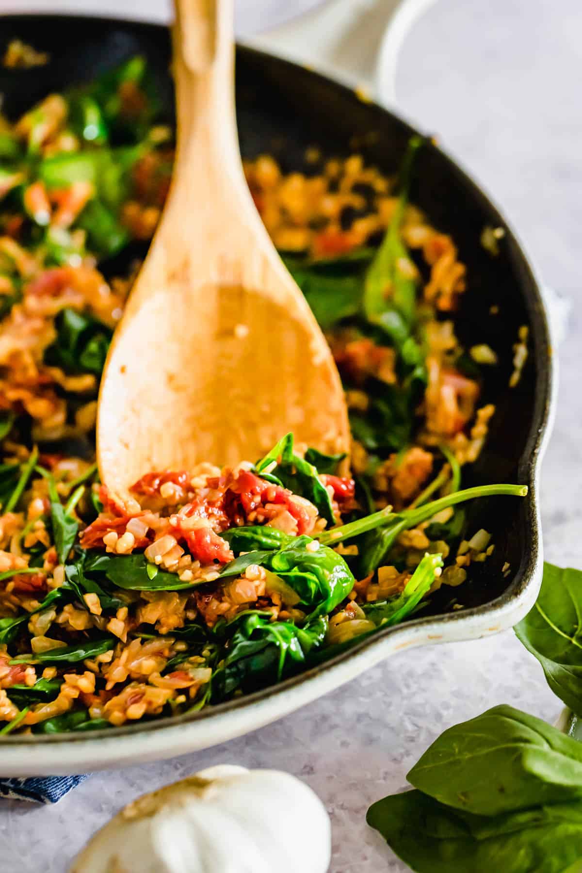 A skillet with spinach and sauteed onion, garlic and sun-dried tomatoes with a wooden spoon, with garlic cloves nearby.