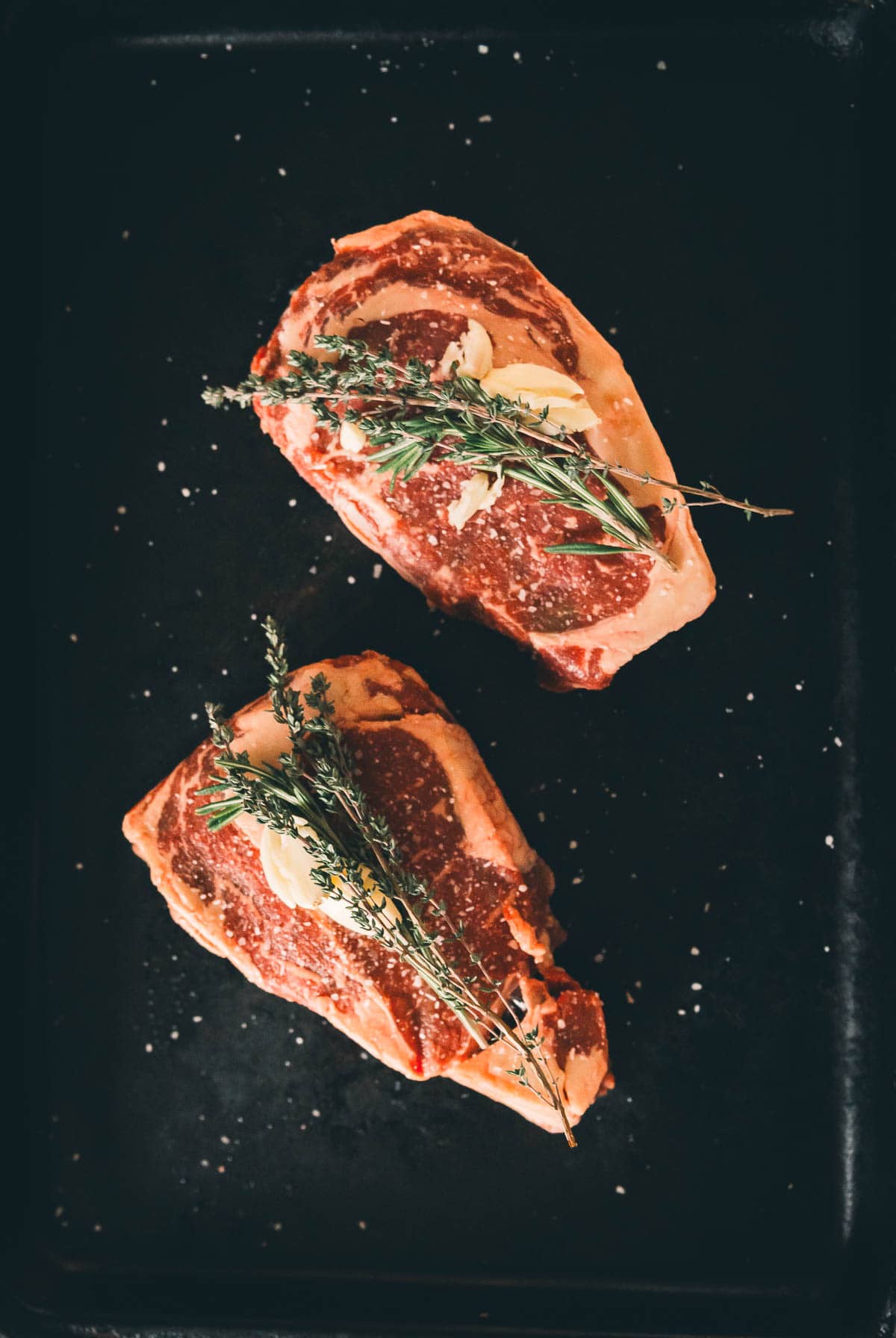 Two raw steaks garnished with garlic and herbs on a dark surface.