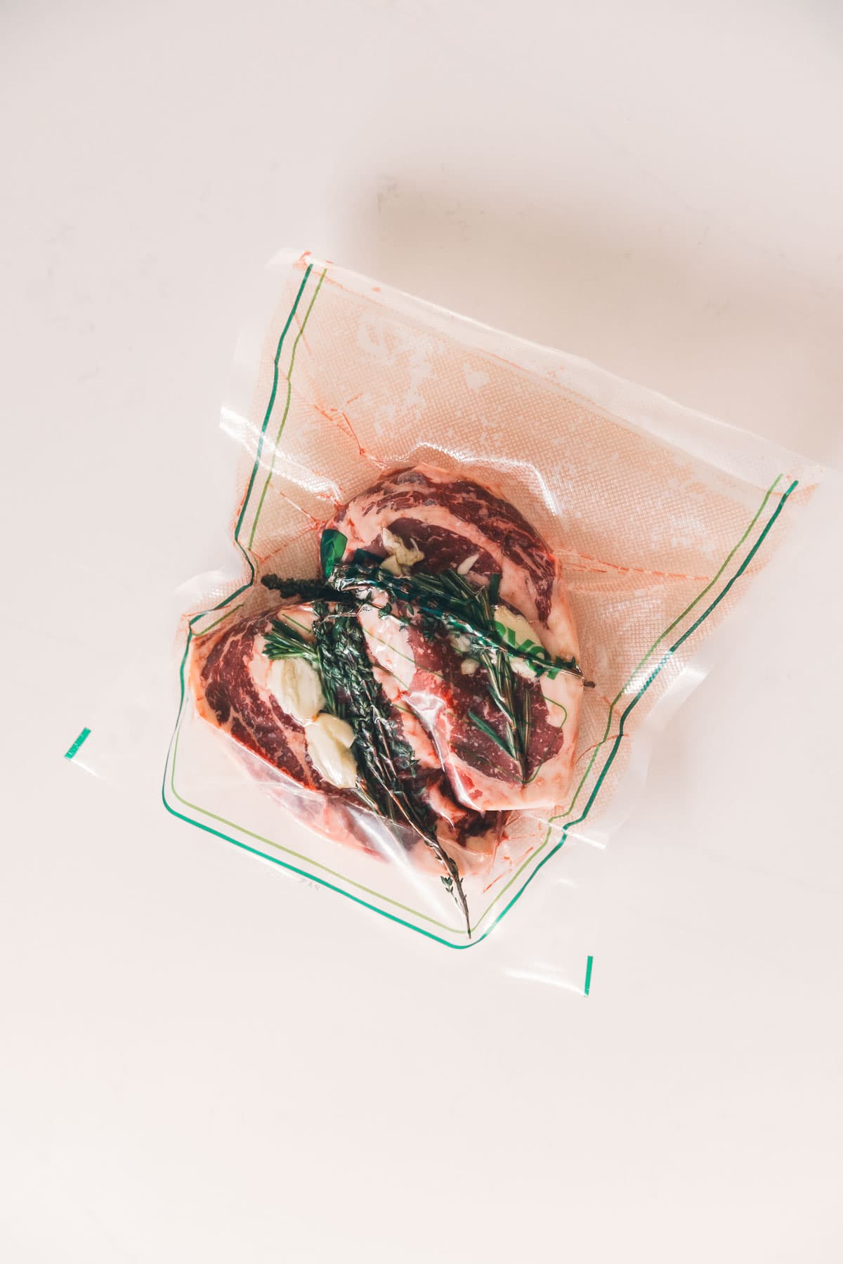 Raw ribeye steaks in a vacuum-sealed bag with herbs and garlic, ready for sous vide cooking, viewed from above on a light background.