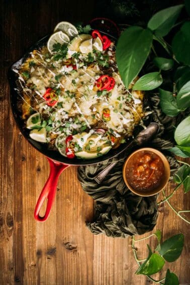 A red skillet filled with nachos garnished with cheese, sliced jalapeños, cilantro, lime, and green onions, accompanied by a side of salsa, on a wooden table surrounded by green leaves.