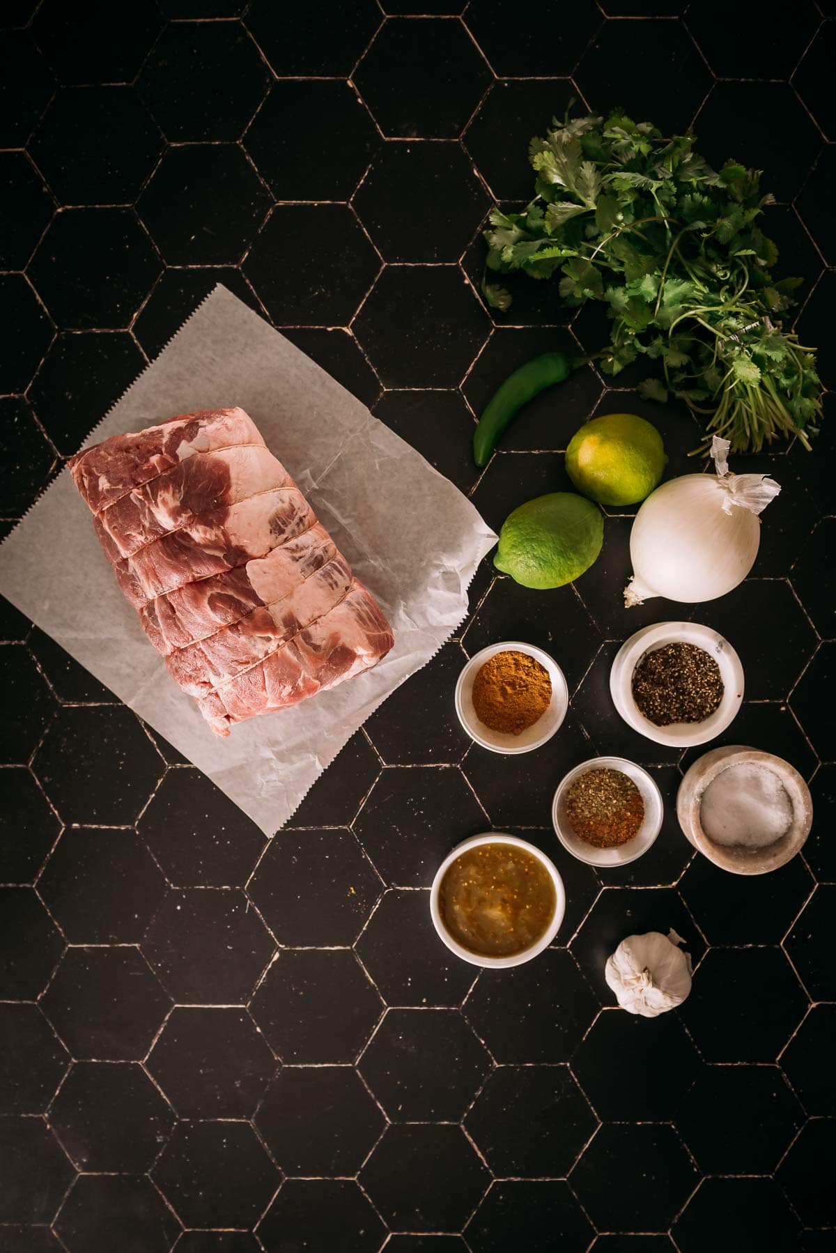 Ingredients for a recipe arranged on a dark tiled surface, including a slab of raw pork shoulder, herbs, spices, lime, and onion.