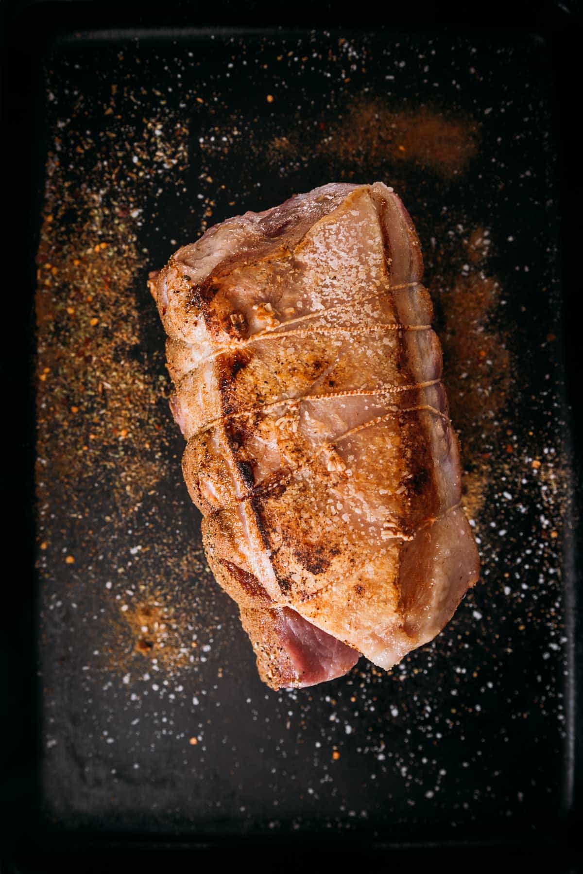 Seared pork fat cap with seasoning on a black tray, viewed from above.