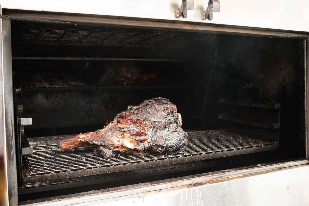 A large steamship round is being smoked inside a metal smoker with a great crust and the bone sticking out.