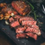 Sliced medium-rare steak served with scallops and herbs on a black plate, garnished with fresh rosemary.