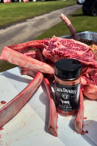 A pile of raw bone-in ribeye steaks is on a table next to a container of Jack Daniel's beef rub seasoning.