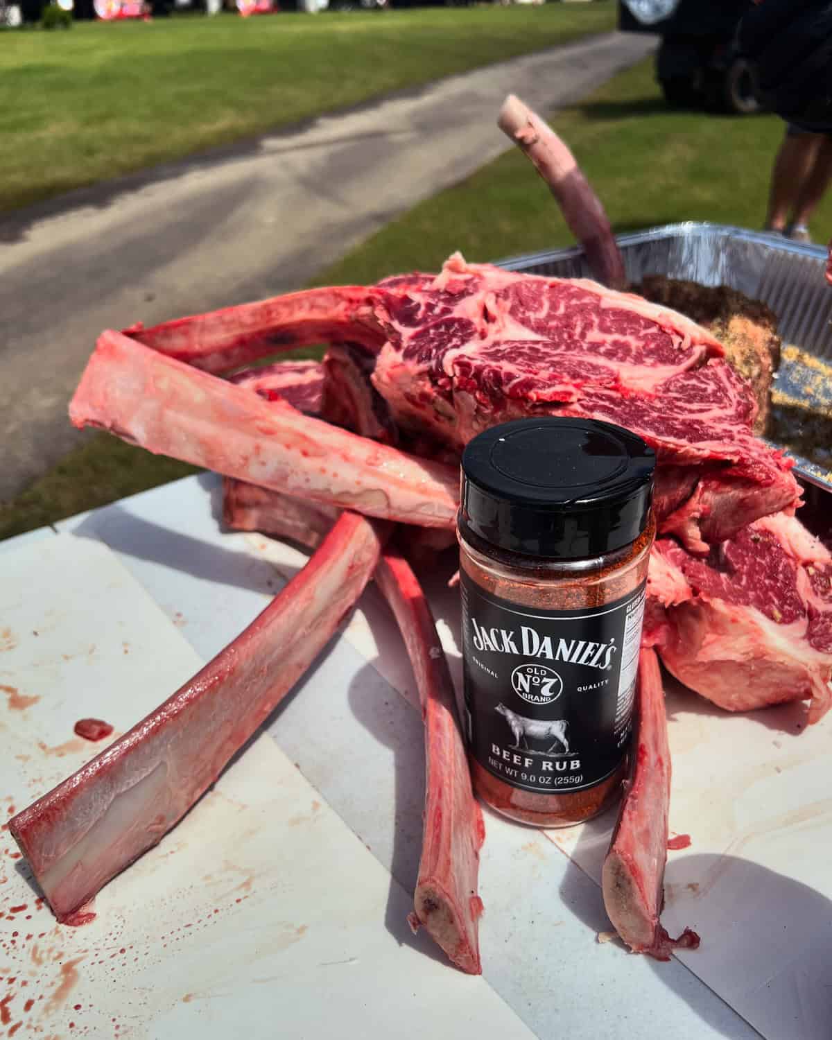 A pile of raw bone-in ribeye steaks is on a table next to a container of Jack Daniel's beef rub seasoning.