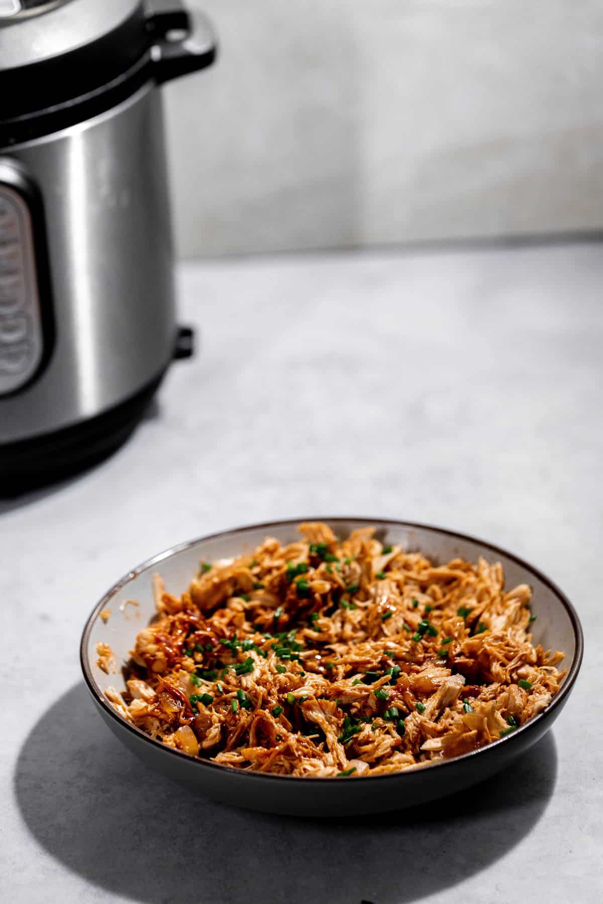 A bowl of shredded chicken garnished with chopped herbs is placed on a countertop, with a pressure cooker in the background.