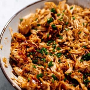 A bowl of shredded chicken topped with finely chopped green herbs and drizzled with a dark sauce.