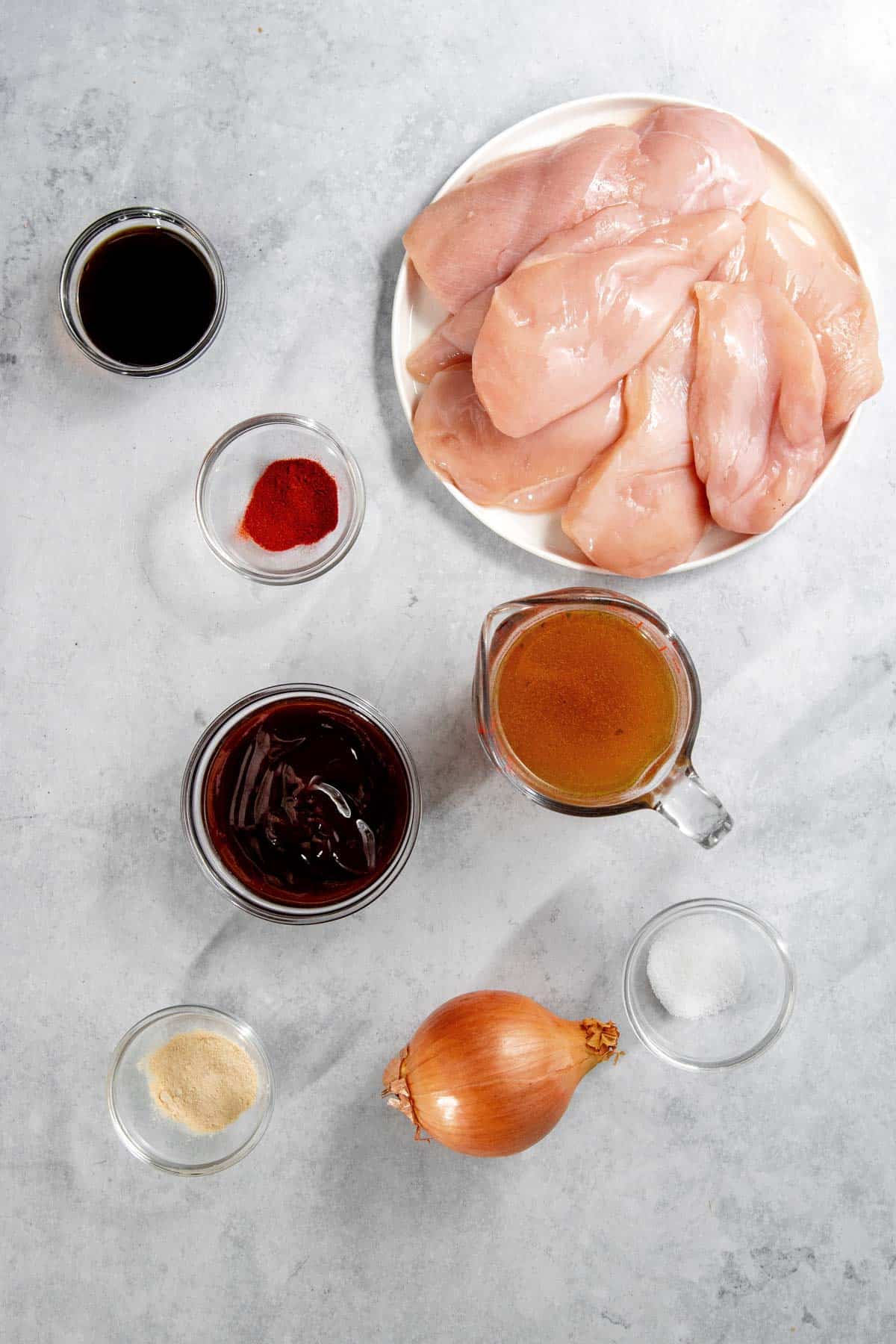 Overhead view of raw chicken breasts on a plate, surrounded by various ingredients in bowls including sauces, spices, onion, and a measuring cup with liquid on a gray surface.