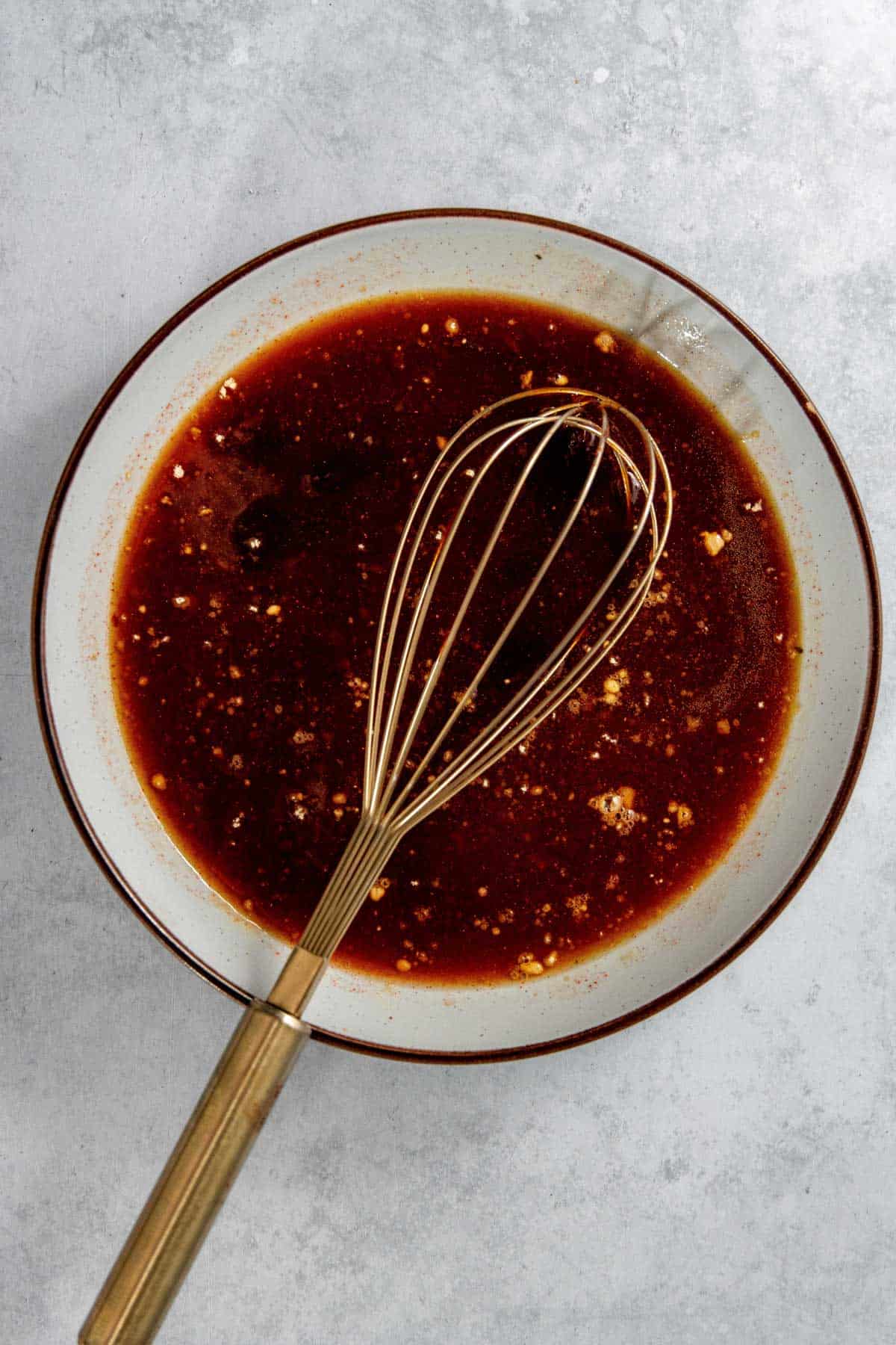 A metal whisk in a bowl filled with a dark brown liquid containing small chunks, on a light gray surface.