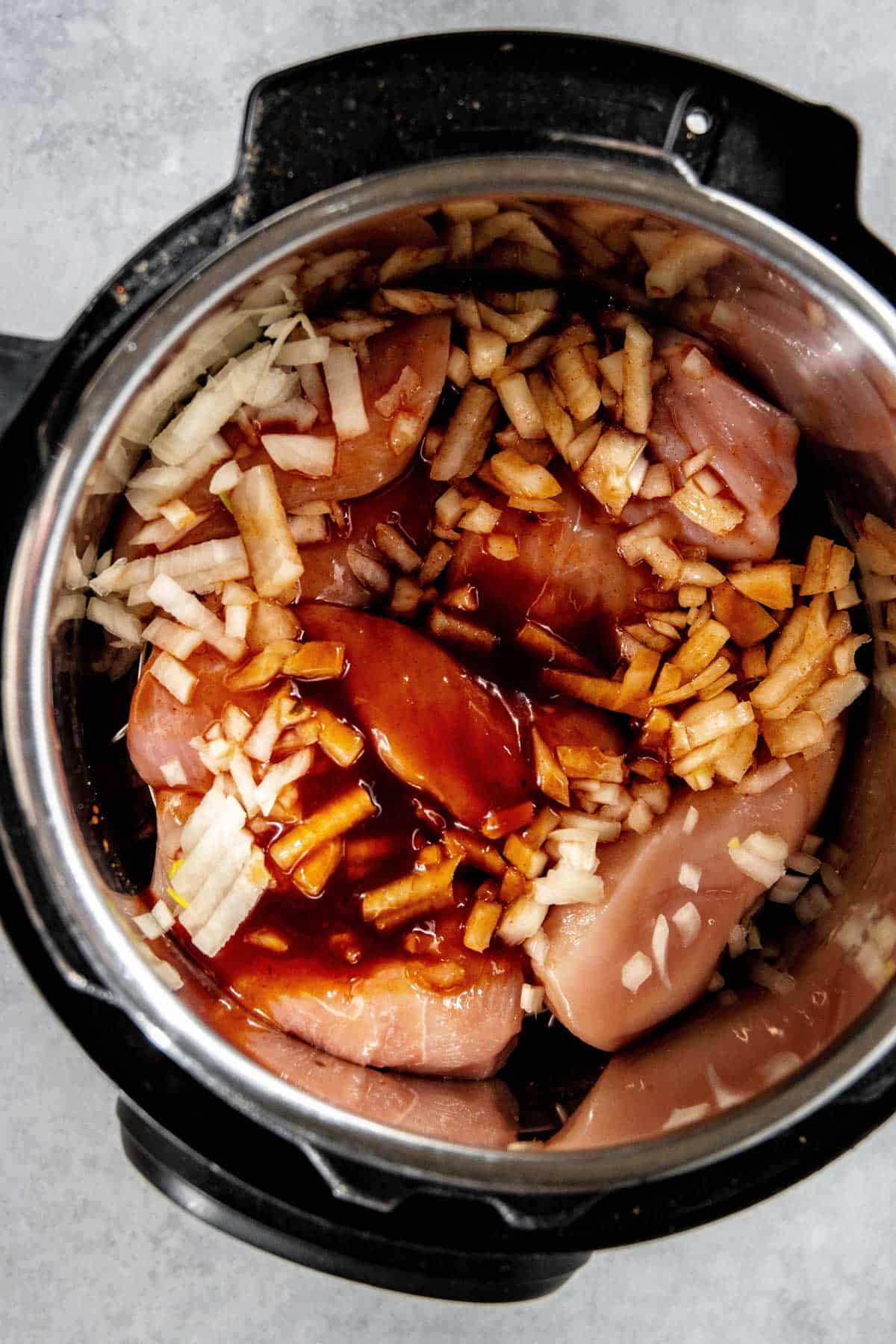 An Instant Pot filled with raw chicken breasts, chopped onions, and a dark liquid sauce.