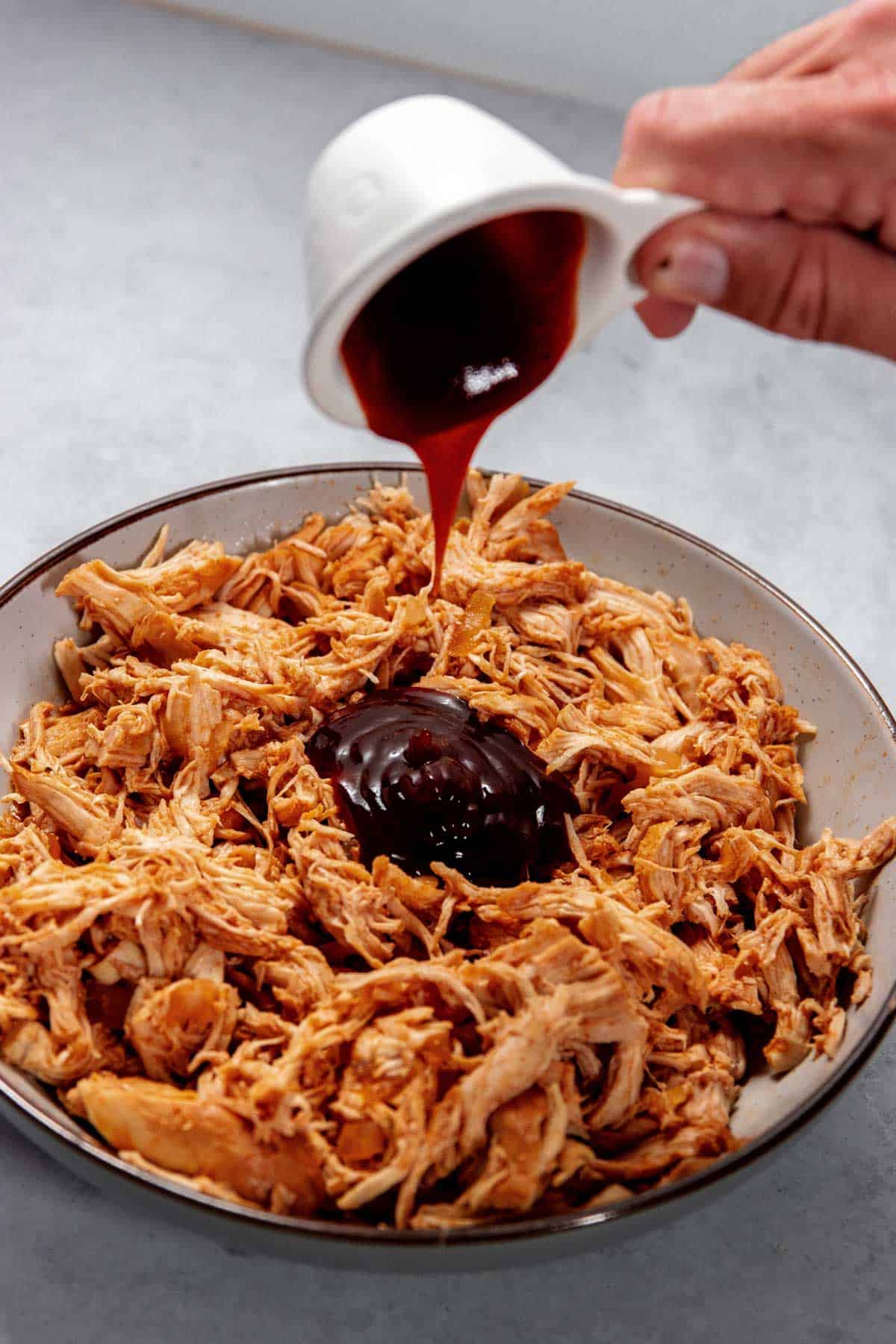 A hand pours barbecue sauce from a white measuring cup onto a bowl of shredded chicken.