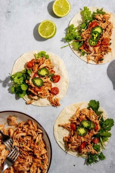 Three chicken tacos with shredded chicken, sliced jalapeños, cherry tomatoes, and cilantro on a white surface. Two lime halves are in the background, and a plate with leftover chicken is on the side.