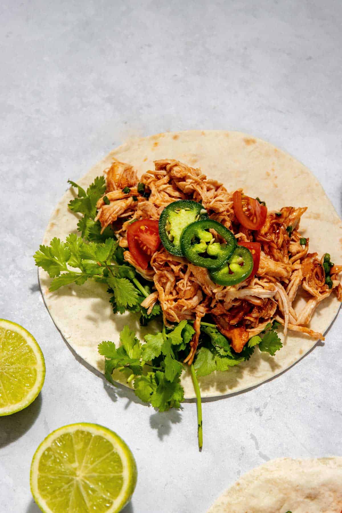 An open taco filled with shredded chicken, cilantro, sliced cherry tomatoes, and jalapeños, resting on a light grey surface. Half lime slices are nearby.