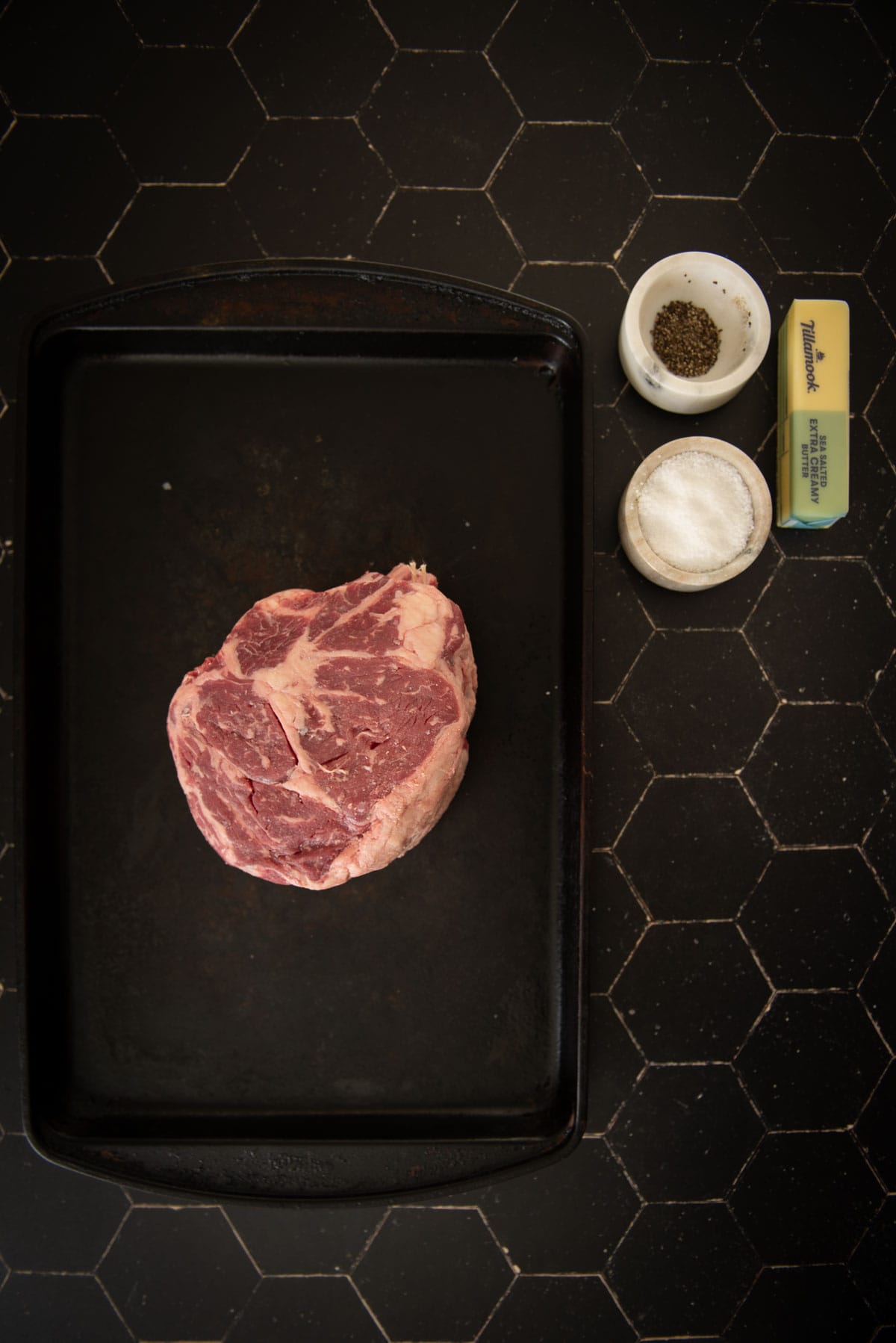 A raw steak on a black baking tray with a cup of pepper, a cup of salt, and a stick of butter on a hexagonal-tiled surface.