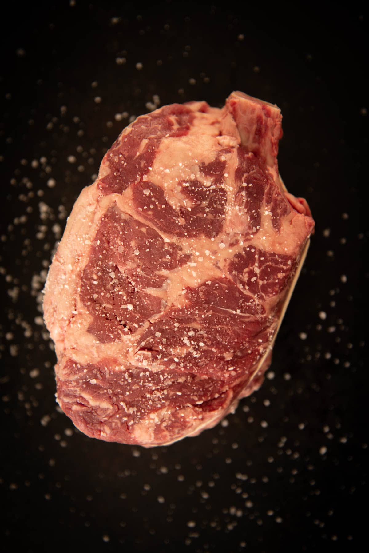 A raw, marbled cowboy steak sprinkled with salt on a dark, speckled surface.