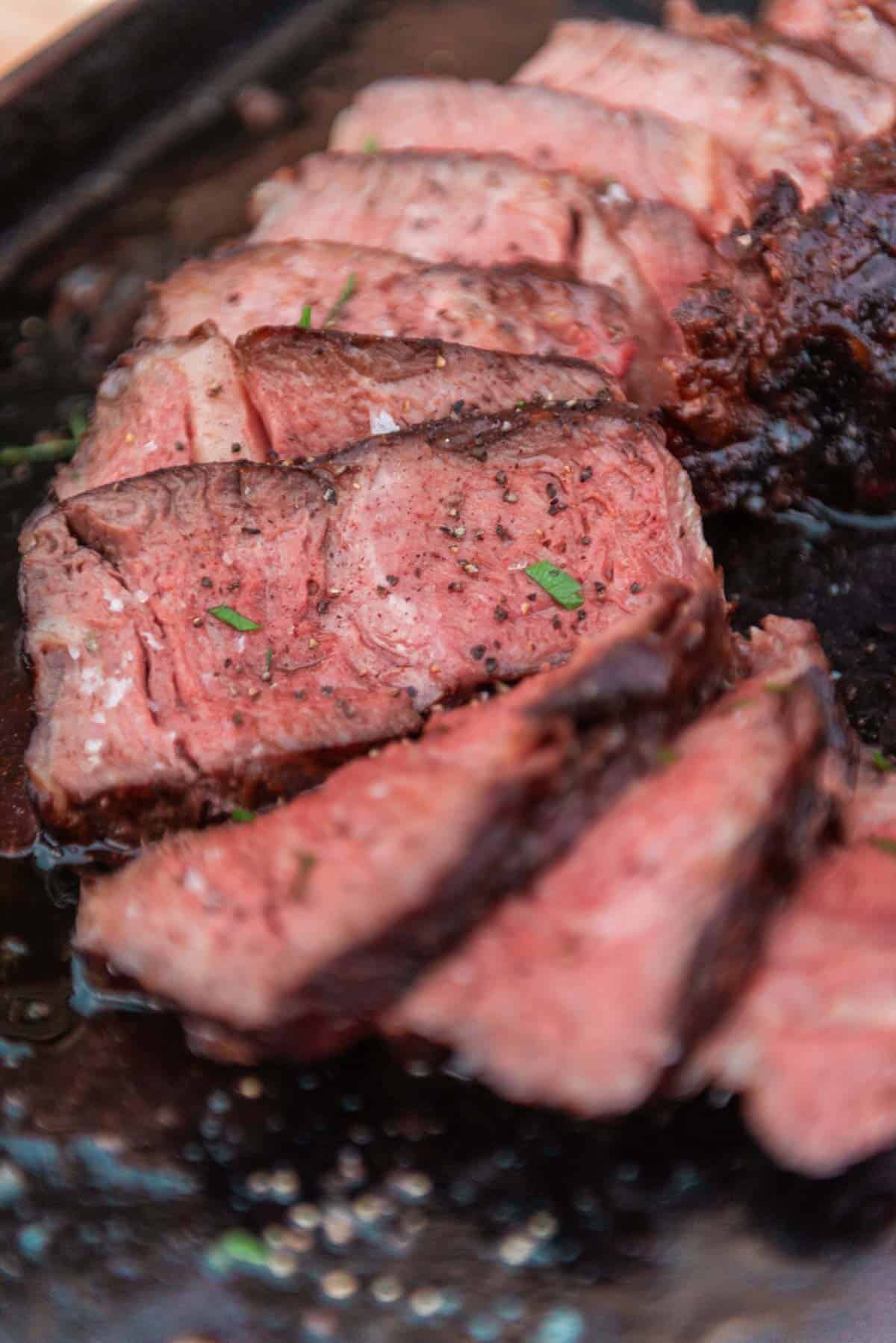 Close-up of sliced, medium-rare cowboy ribeye seasoned with herbs and pepper on a black plate.