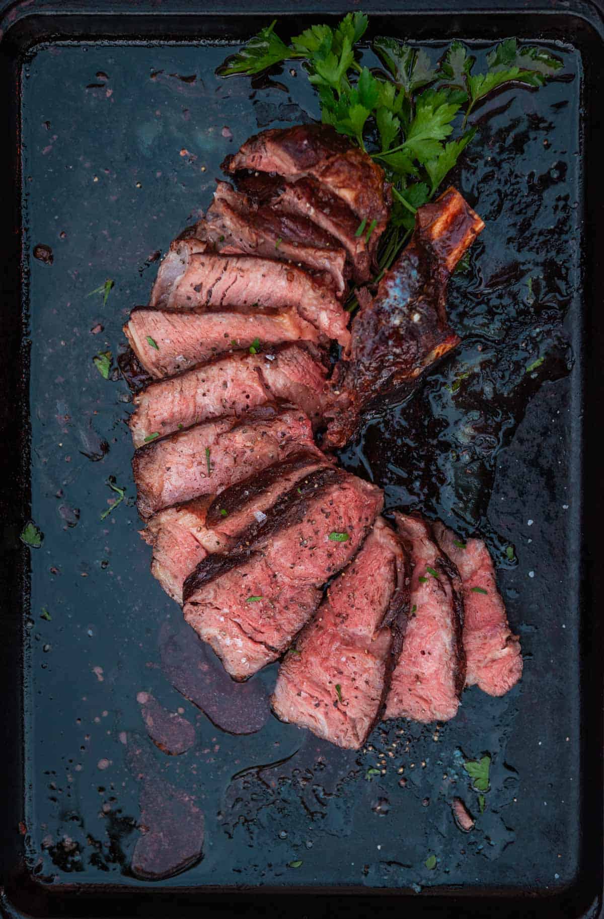 Sliced, smoked cowboy steak, sliced and arranged in a fanned-out pattern on a dark surface with greens as garnish.