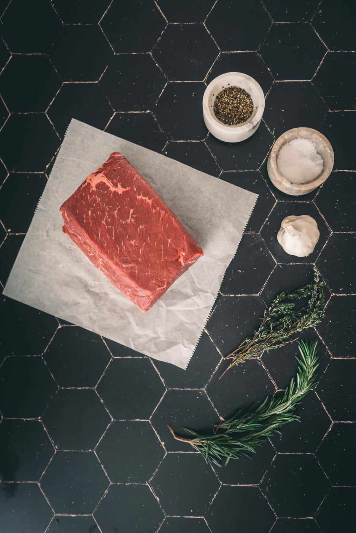 A raw bottom round roast on parchment paper, next to a garlic bulb, herbs, a small bowl of salt, and a small bowl of pepper, arranged on a black hexagon-tiled surface.