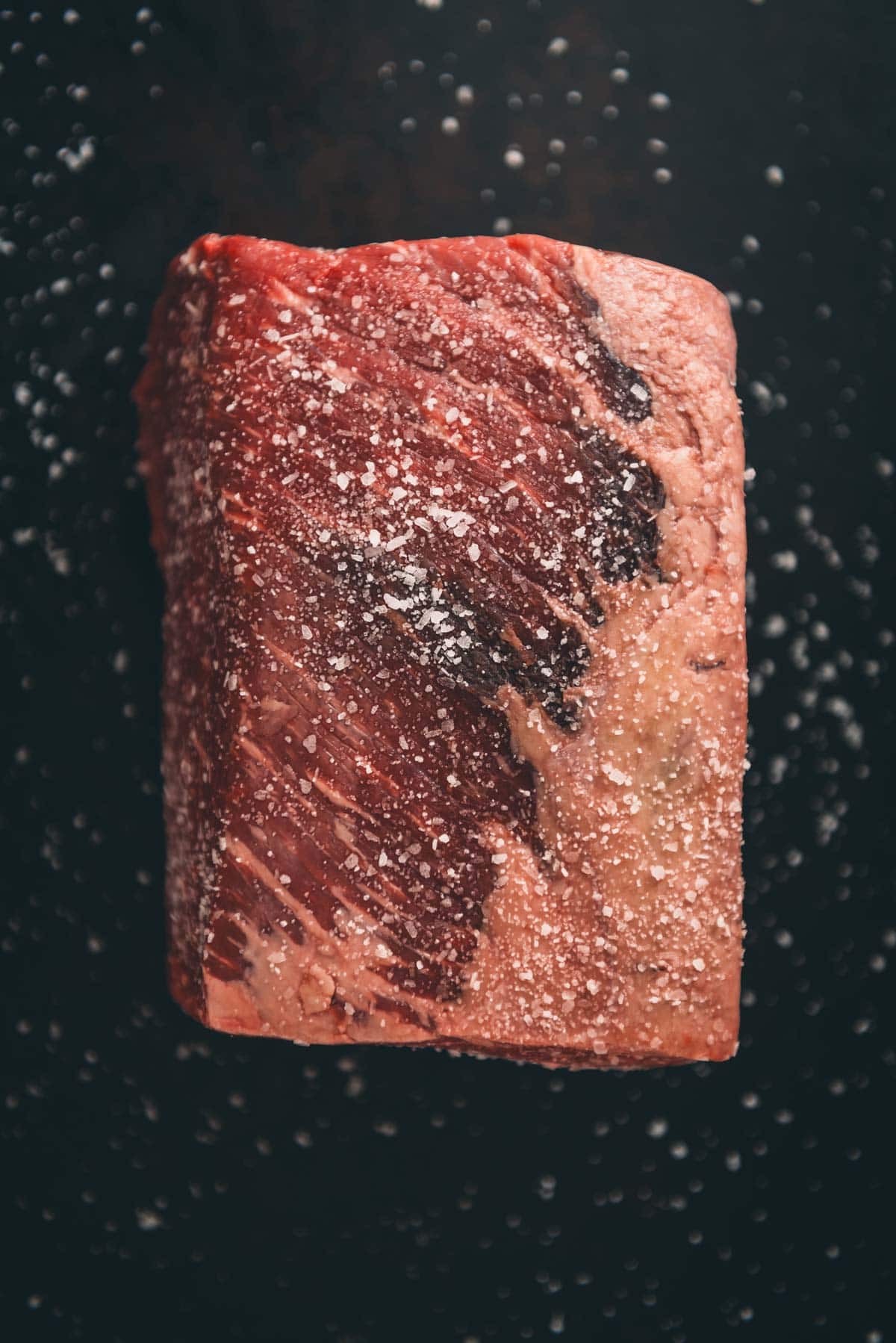 A raw round roast covered in coarse salt is set against a dark background.