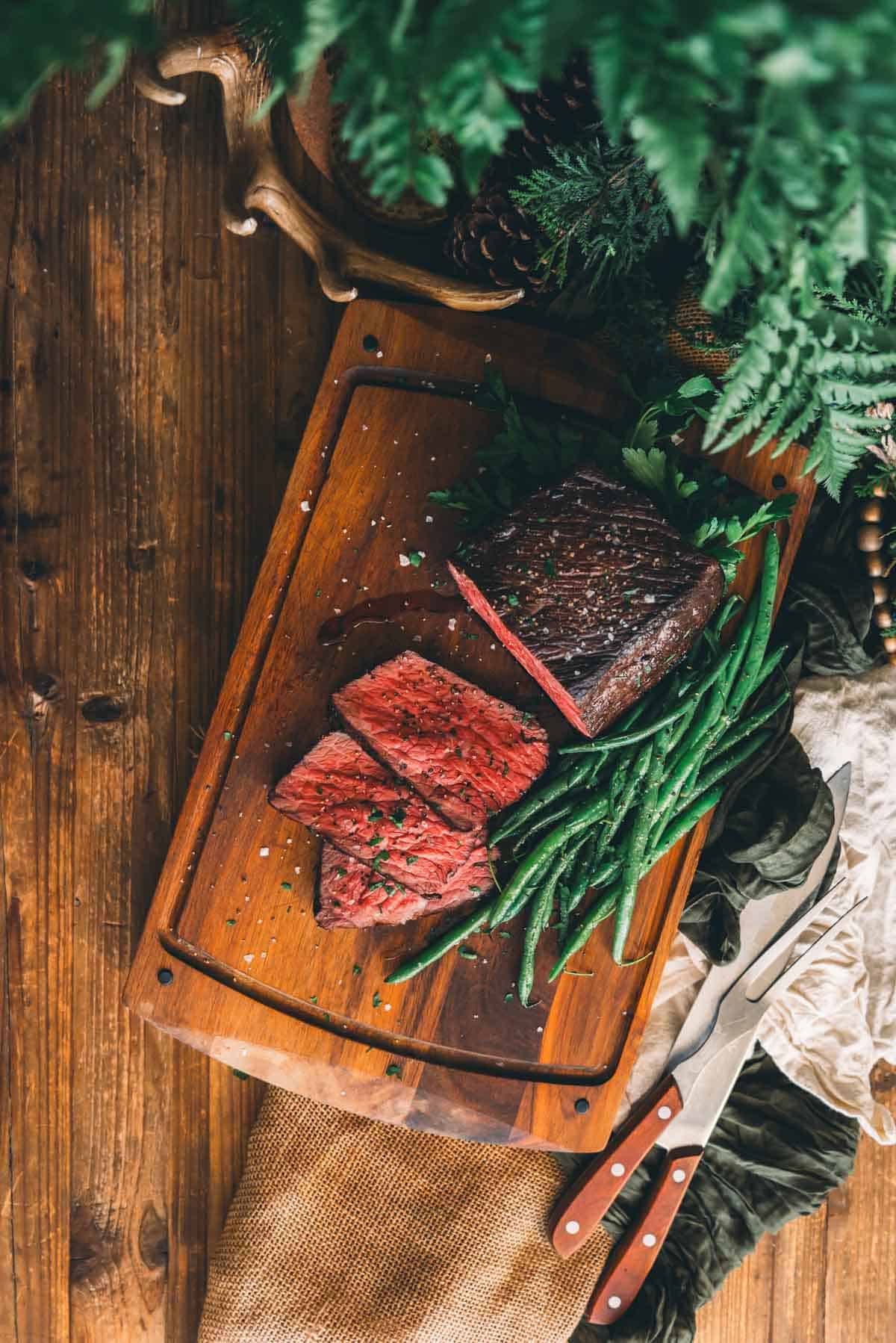 A wooden cutting board with sous vide roast beef carved into slices and green beans, accompanied by a knife and fork on a rustic wood table with greenery.