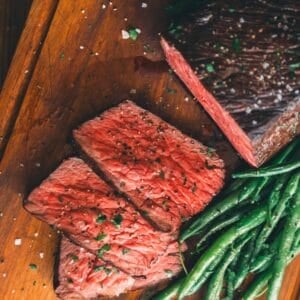 Sliced medium-rare roast beef garnished with herbs, accompanied by a serving of green beans on a wooden cutting board.