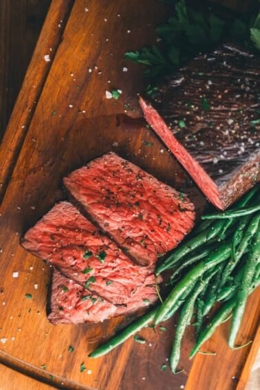 Sliced medium-rare roast beef garnished with herbs, accompanied by a serving of green beans on a wooden cutting board.