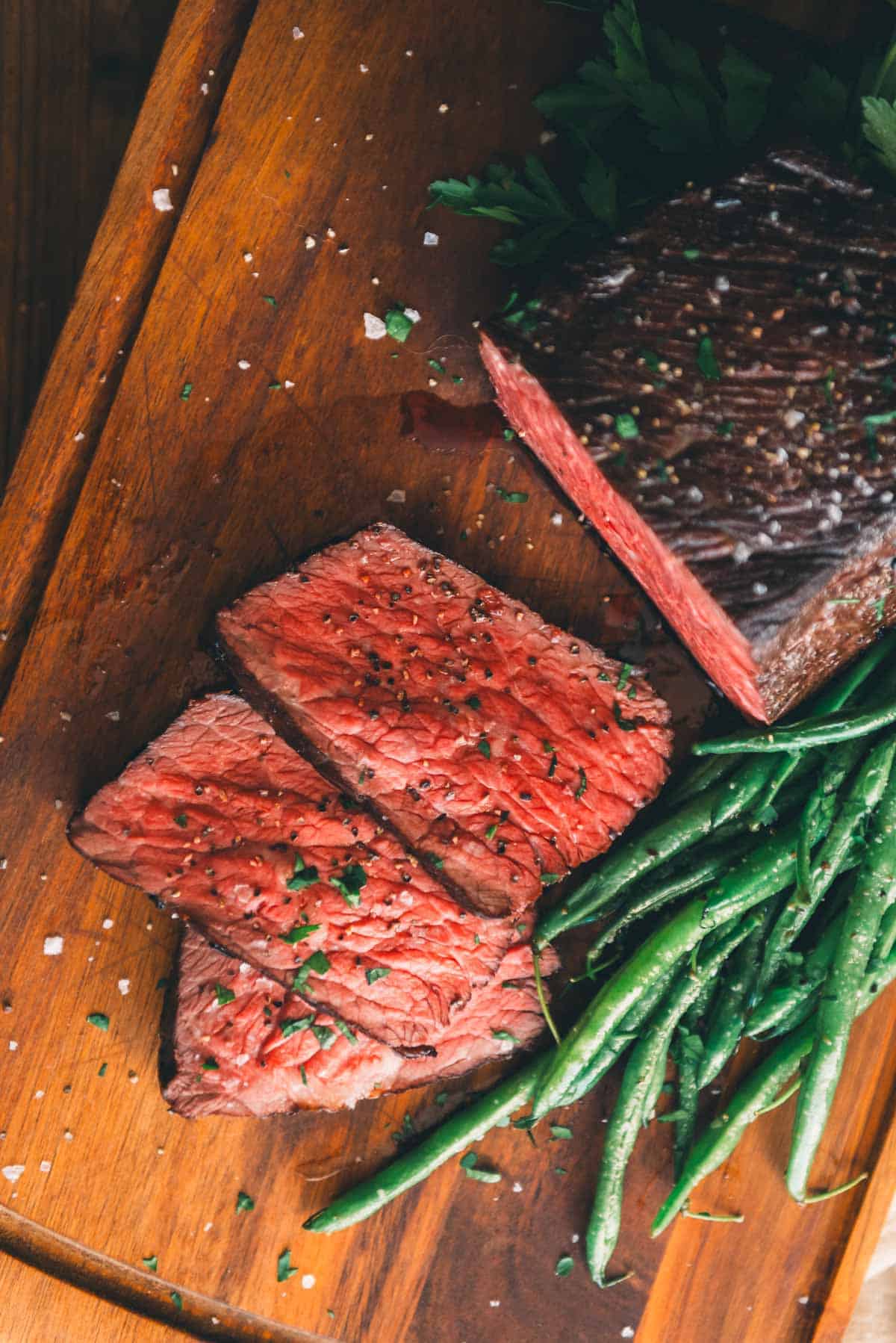 Sliced medium-rare roast beef garnished with herbs, accompanied by a serving of green beans on a wooden cutting board.