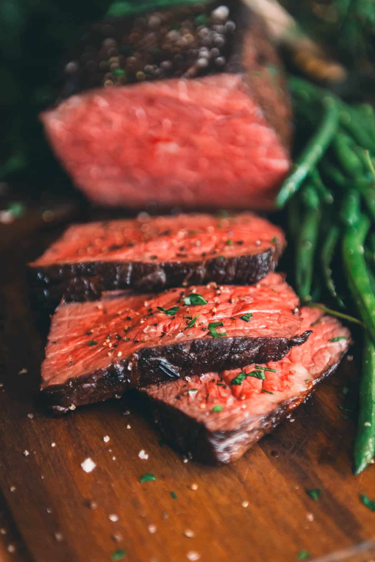 Close-up of sliced medium-rare roast beef garnished with herbs, placed on a wooden board with green beans on the side.