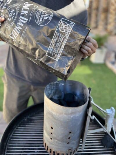 Person pouring charcoal from a Jack Daniel's bag into a charcoal chimney starter on a barbecue grill.
