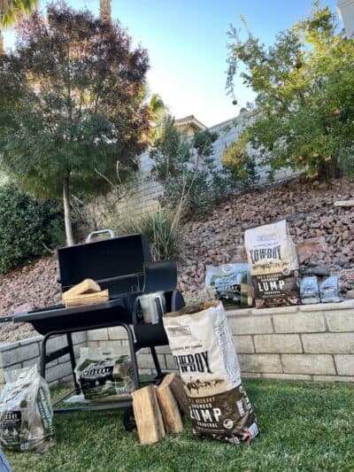 A charcoal grill in a backyard is surrounded by various bags of Cowboy brand hardwood lump charcoal and wood chunks.