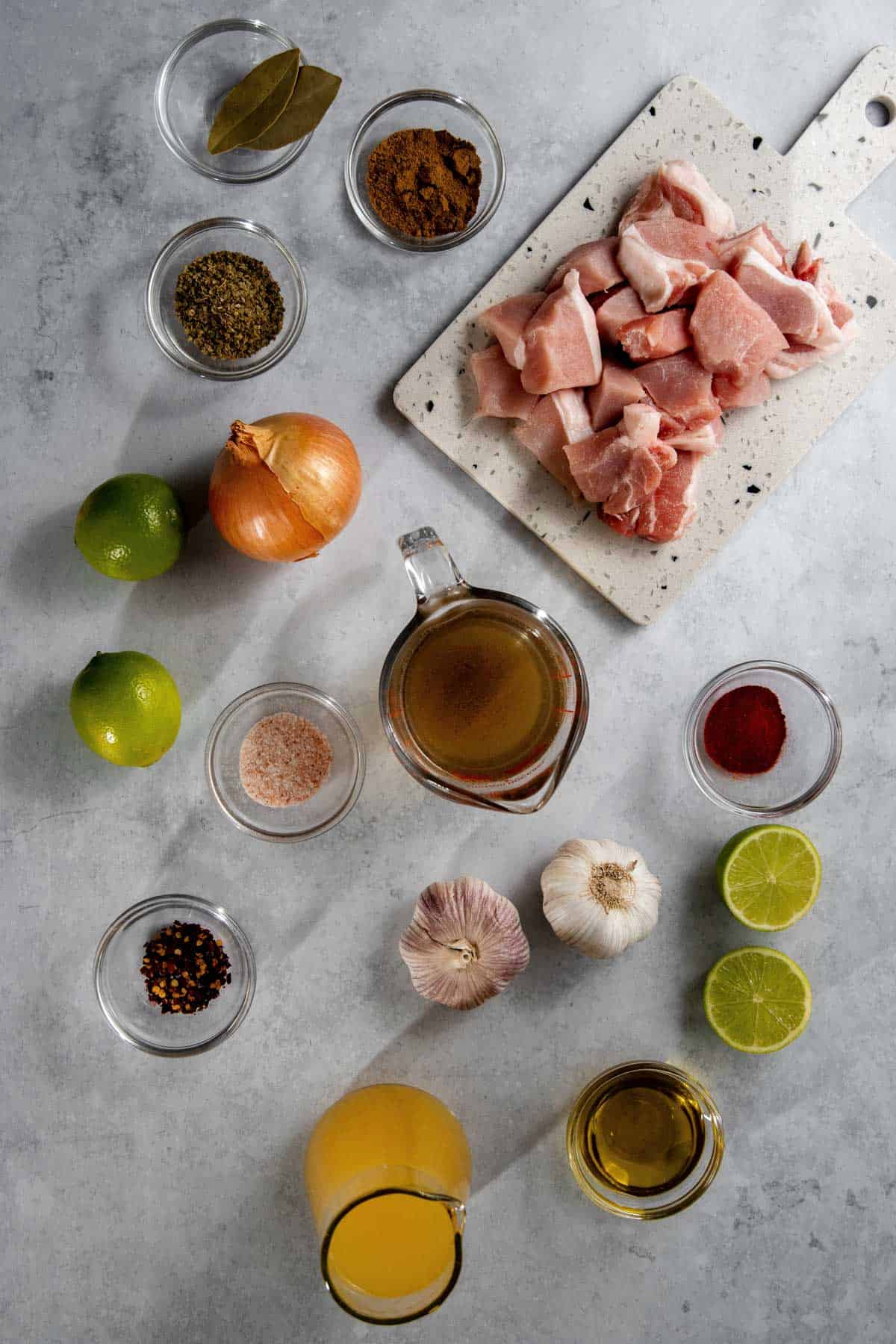 Top-down view of cooking ingredients including cubed raw pork, spices, lime, onion, broth, olive oil, garlic, and cutting board arranged on a gray surface.