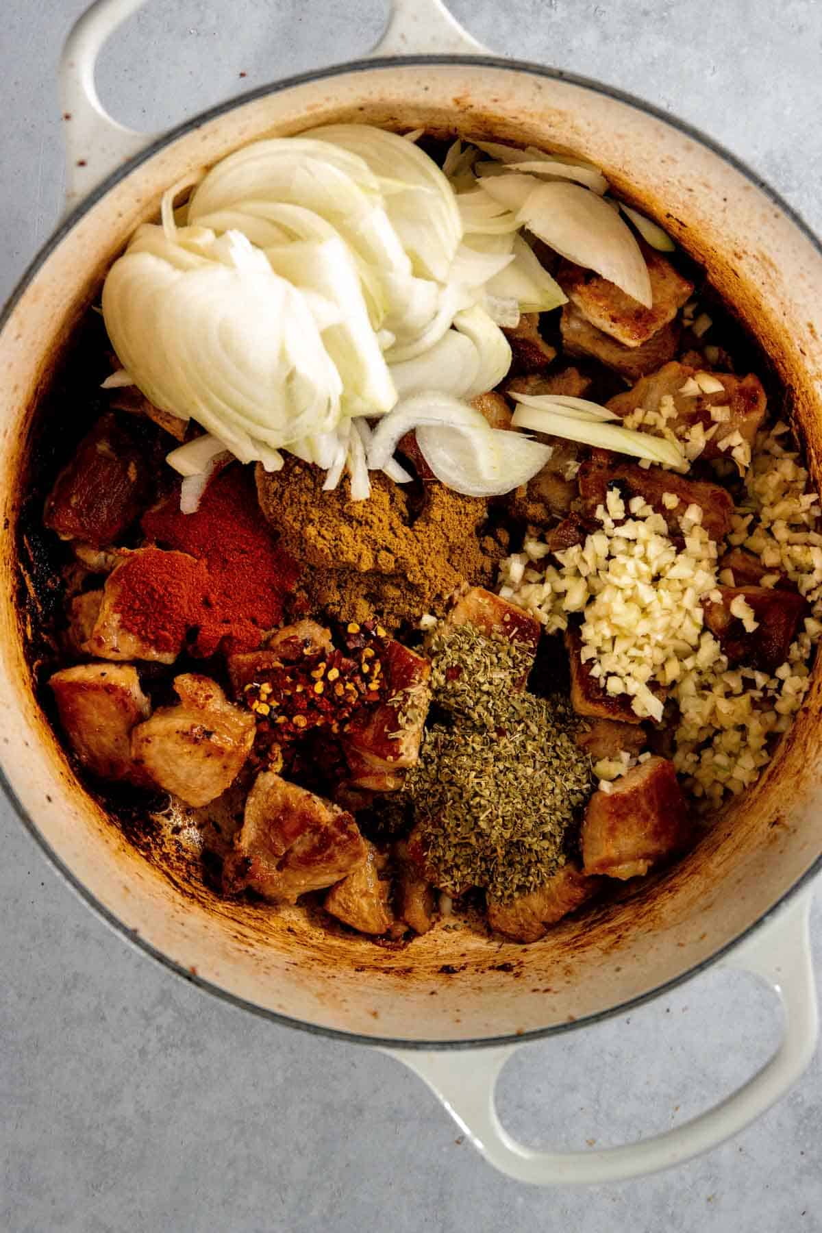 A pot filled with cooked meat pork, sliced onions, minced garlic, and various spices like paprika and oregano on a gray surface.